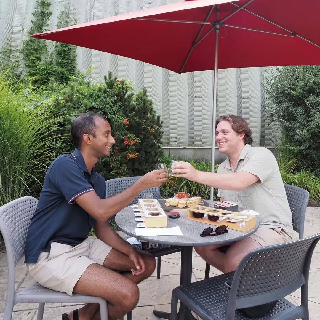 Two guests raising wine glasses at a table with a baked dip and baguette, vineyard rows behind